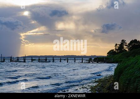 La lumière dorée du soleil jette un œil à travers les nuages orageux tandis que les vagues tapent doucement contre le rivage près d'une ancienne jetée en bois, créant une atmosphère tranquille mais dramatique. Banque D'Images