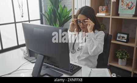 Jeune femme hispanique, médecin en clinique, brune portant des lunettes, travaille sur ordinateur au bureau, ajustant les lunettes. Banque D'Images