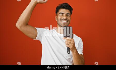 Souriant jeune homme hispanique avec la barbe chantant à l'aide d'une brosse à cheveux sur fond rouge isolé Banque D'Images