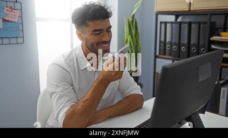 Jeune homme dans un bureau envoyant un message vocal sur son smartphone tout en souriant et assis à un bureau avec un écran d'ordinateur et des fournitures de bureau Banque D'Images