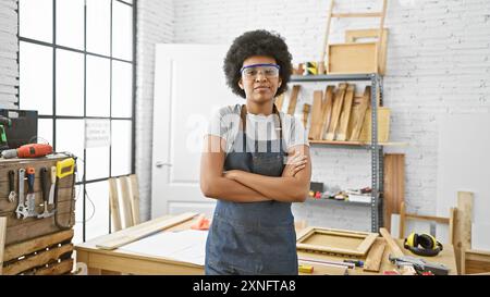 Femme noire confiante avec les cheveux bouclés portant des lunettes de sécurité se croise les bras dans un atelier de menuiserie intérieure. Banque D'Images