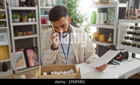 Jeune homme avec la barbe parlant au téléphone tout en examinant des documents dans un magasin de décoration d'intérieur entouré de divers articles de décoration et de plantes Banque D'Images