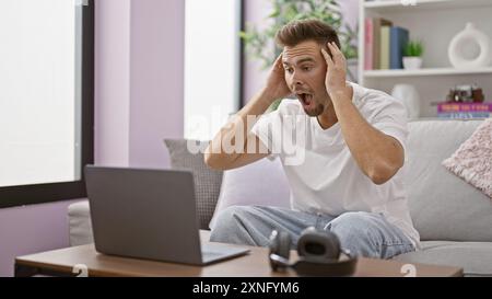 Choqué jeune homme hispanique avec la barbe dans le salon réagissant au contenu de l'écran d'ordinateur portable à la maison. Banque D'Images