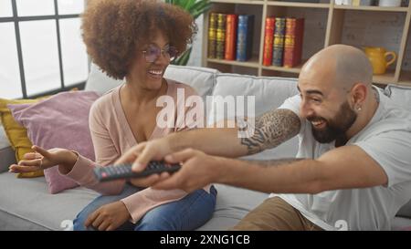 Couple interracial ludique combattant sur la télécommande de télévision dans un salon confortable. Banque D'Images