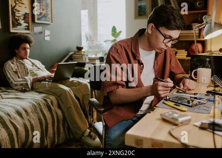 Jeune technicien soudant de petits détails d'ordinateur tandis que son ami travaille à distance sur le lit en arrière-plan Banque D'Images