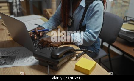 Une femme afro-américaine travaillant comme détective tape sur un ordinateur portable dans un bureau de poste de police. Banque D'Images