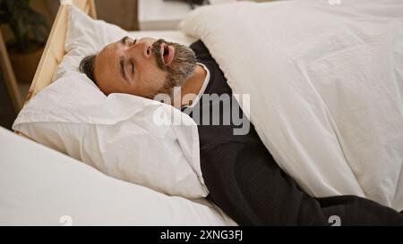 Bel homme hispanique senior avec une barbe grise dormant paisiblement dans une chambre à coucher à la maison. Banque D'Images