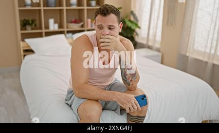 Homme hispanique pensif avec des tatouages est assis sur le lit tenant un smartphone dans un intérieur de chambre minimaliste. Banque D'Images