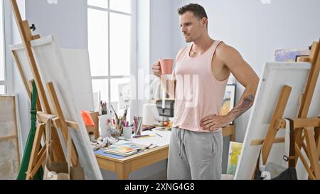 Bel homme tenant une tasse dans un studio d'art ensoleillé avec chevalet et fournitures de peinture Banque D'Images