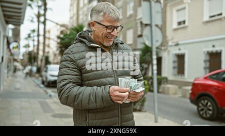 L'homme aux cheveux gris souriant compte les dollars américains dans une rue urbaine animée, illustrant la finance dans la vie quotidienne. Banque D'Images