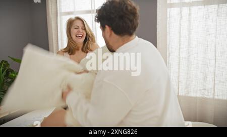 Une femme et un homme joyeux s'engagent dans un combat d'oreillers ludique dans une chambre ensoleillée, incarnant l'amour et le bonheur ensemble dans un cadre domestique. Banque D'Images