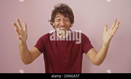 Un jeune homme hispanique joyeux avec une barbe, portant une chemise marron décontractée, pose les bras ouverts contre un mur rose Uni. Banque D'Images