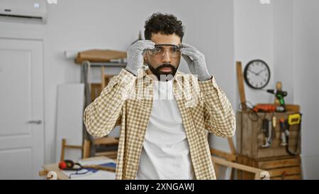 Beau jeune homme avec la barbe mettant des lunettes de sécurité dans une salle d'atelier de menuiserie. Banque D'Images