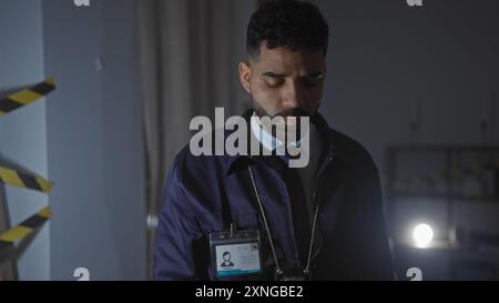 Le détective hispanique analyse les preuves à l'intérieur sur une scène de crime faiblement éclairée, paraissant pensif et concentré. Banque D'Images