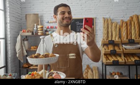 Jeune homme hispanique souriant dans une boulangerie tout en prenant un selfie sur un smartphone, entouré de pain et de viennoiseries Banque D'Images