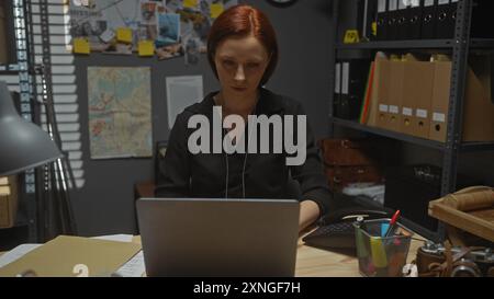 Une femme rousse concentrée travaille sur son ordinateur portable dans le bureau d'un détective, entourée de preuves et de dossiers. Banque D'Images