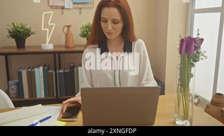 Une femme rousse concentrée utilise un ordinateur portable à la maison, un smartphone à la main, suggérant un style de vie professionnel moderne. Banque D'Images