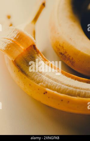 Deux bananes jaunes reposent sur une table blanche, baignées de lumière chaude du soleil. L'un d'eux a sa peau partiellement enlevée. Les fruits sont des aliments sains. Banque D'Images