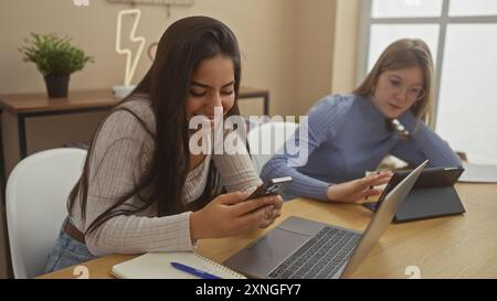 Deux femmes utilisant des appareils technologiques tout en étant assises ensemble dans un environnement de bureau à domicile moderne, affichant un travail décontracté et la communication Banque D'Images