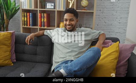 Bel homme noir se relaxant sur un canapé dans un salon confortable tout en tenant une télécommande. Banque D'Images