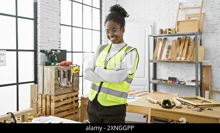 Une femme africaine confiante, les bras croisés, se tient debout dans un atelier de menuiserie bien équipé, mettant en valeur la diversité au travail. Banque D'Images