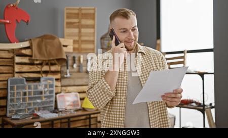 Bel homme caucasien parlant au téléphone dans un atelier de menuiserie, inspectant un document. Banque D'Images