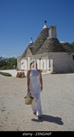 Une jeune femme hispanique en vêtements blancs marche avec un sac de paille devant les maisons traditionnelles de trulli dans la vieille ville d'alberobello, pouilles, italie, au Nouveau-Brunswick Banque D'Images