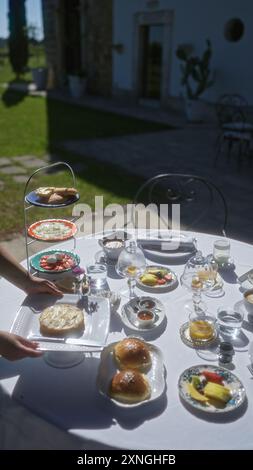 La main d'une femme place une assiette sur une table de petit-déjeuner bien décorée à l'extérieur dans une villa italienne dans les pouilles, en europe, mettant en valeur une étendue élégante du passé Banque D'Images