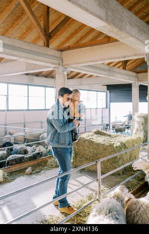 Papa souriant avec petite fille dans ses bras regardant des moutons dans le paddock à la ferme Banque D'Images