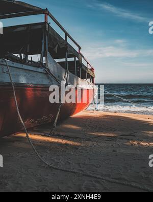 Un bateau en bois rouge et blanc repose sur une plage de sable. L'eau calme et le ciel clair créent une scène paisible. Banque D'Images