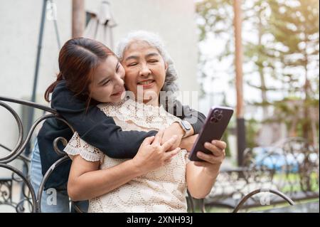 Une jeune petite-fille asiatique attentionnée et heureuse serre sa grand-mère dans ses bras pendant qu'elle utilise son téléphone sur un banc dans un jardin. liens familiaux, ensemble, amour, Banque D'Images