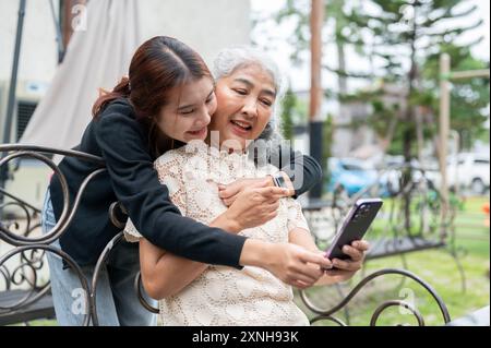 Une jeune petite-fille asiatique attentionnée et heureuse serre sa grand-mère dans ses bras pendant qu'elle utilise son téléphone sur un banc dans un jardin. liens familiaux, ensemble, amour, Banque D'Images