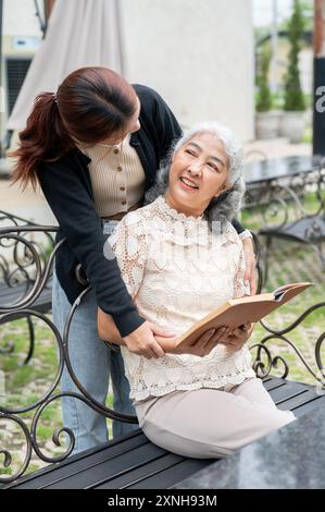 Une jeune petite-fille asiatique attentionnée et heureuse embrasse sa grand-mère pendant qu'elle lit un livre sur un banc dans un jardin. liens familiaux, ensemble, amour, gr Banque D'Images
