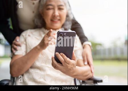 Une image en gros plan d'une heureuse grand-mère asiatique retraitée utilisant un smartphone assise dans un fauteuil roulant, avec son soignant ou sa fille la promenant dans le p Banque D'Images