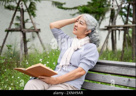 Une femme asiatique mature de 60 ans touche sa tête, ressentant un mal de tête et des étourdissements, en lisant un livre dans son jardin. problème de soins de santé Banque D'Images