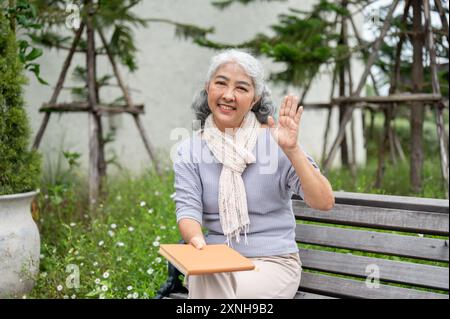 Une heureuse femme asiatique retraitée en vêtements décontractés agite sa main et sourit à la caméra tout en étant assise sur un banc dans son jardin, tenant un bo Banque D'Images