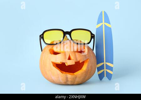 Citrouille sculptée pour Halloween avec des lunettes de soleil élégantes et planche de surf sur fond bleu Banque D'Images