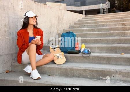 Jeune femme asiatique dans des écouteurs avec des boîtes de peinture en aérosol à l'aide d'un téléphone portable dans les escaliers à l'extérieur Banque D'Images