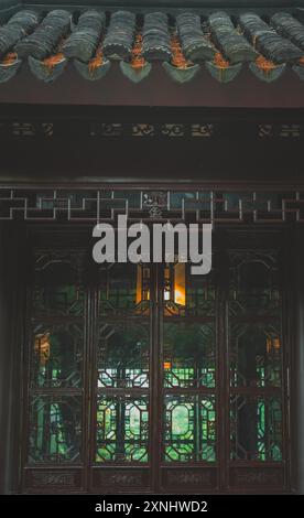 Vue sur un jardin chinois traditionnel. Une porte en bois avec des sculptures complexes offre un aperçu de la végétation luxuriante. Banque D'Images