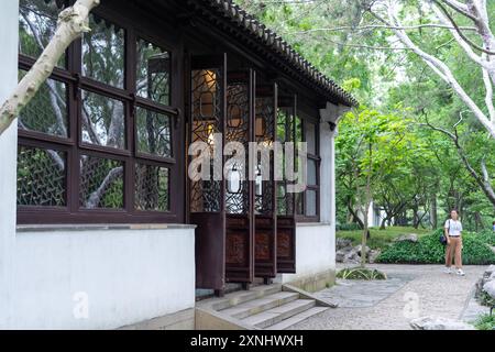 Suzhou, Chine - 11 juin 2024 : Un bâtiment de jardin chinois traditionnel avec un mur blanc et une porte en bois avec des sculptures complexes, entouré de luxuriants Banque D'Images