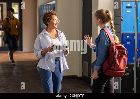 Parlant dans le couloir du lycée, deux adolescentes avec tablette et sac à dos près des casiers Banque D'Images