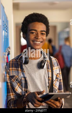 Adolescent souriant tenant une tablette et portant des écouteurs dans le couloir du lycée Banque D'Images