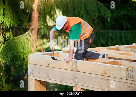 Menuisier construisant une maison à ossature en bois de deux étages près de la forêt. Homme martelant des clous dans la structure tout en portant un casque de protection et un gilet de construction. Concept de construction écologique moderne Banque D'Images