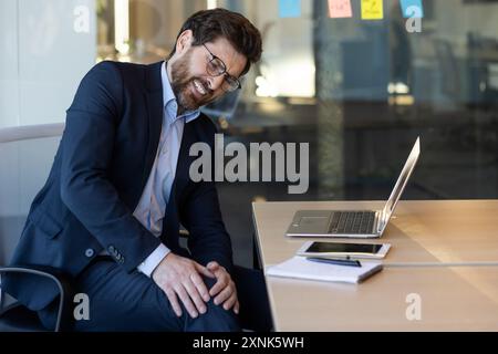 Homme d'affaires en tenue formelle éprouvant des douleurs à la jambe alors qu'il était assis au bureau dans l'environnement de bureau. L'homme tient le genou dans l'inconfort, ordinateur portable et bloc-notes sur le bureau, fond d'espace de travail moderne. Banque D'Images