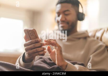Homme heureux dans le sweat à capuche brun en utilisant le téléphone, étudier, apprendre, faire des achats, travailler en ligne, parler, écouter de la musique, livre audio, podcast tout en étant assis Banque D'Images