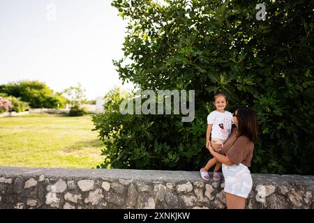 Mère tenant sa fille souriante dans un jardin extérieur ensoleillé, capturant un moment de famille heureux pendant une journée d'été. Banque D'Images