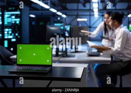 Gros plan de l'ordinateur portable à écran vert dans le centre de données avec les travailleurs vérifiant les documents des procédures d'urgence. Concentrez-vous sur l'ordinateur portable Chroma Key devant les employés du hub de serveurs en arrière-plan flou Banque D'Images