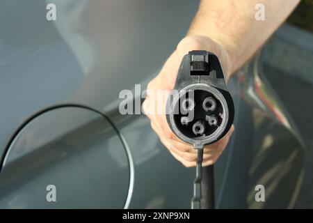 Homme tenant le câble d'alimentation près de la voiture à la station de charge de véhicule électrique, gros plan Banque D'Images