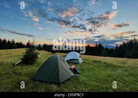 Peu de tentes touristiques dans un champ herbeux le soir, entourées d'arbres et de collines sous le ciel magnifique du coucher du soleil. Les nuages colorés rehaussent la beauté de la scène, avec des teintes orange et violet illuminant l'horizon. Banque D'Images