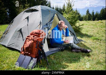 L'homme est assis dans l'entrée de la tente touristique, étudiant la carte avec une expression réfléchie. À côté de lui sac à dos orange, équipé d'un trépied et d'un panneau solaire, placé sur fond pittoresque d'arbres et de montagnes. Banque D'Images